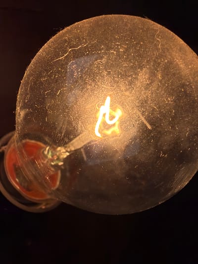 A close-up, top-down view captures the filament of an incandescent light bulb, glowing warmly against a dark background. The glass bulb is coated in a fine layer of dust and appears slightly textured. The filament itself, a bright, twisted wire, emits a strong yellow-orange light. At the base of the bulb, a red component is visible within the clear glass housing. The scene is indoors, likely a home, in the village of Papyarnyanski Sel'ski Savet, Belarus, as there are no external environmental factors visible.