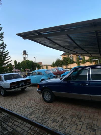 The image shows a row of parked cars under a covered structure, possibly a carport. The cars are of various colors, including blue, light blue, maroon, and white. A water tower is visible in the background. The location is Kwang, Nigeria.