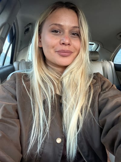 The image depicts a woman with long, blonde hair in the front passenger seat of a car. She is wearing a brown jacket. The interior of the car is visible, including the dashboard, seats, and window. The location in Дулесово, Russia, cannot be confirmed from the image.