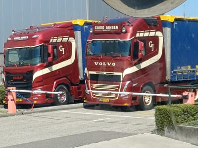 In Venlo, Netherlands, two large red transport trucks are parked in front of a modern building with a grey, corrugated metal facade. The truck on the left is a DAF, and the truck on the right is a Volvo. Both trucks have "GUIDO JANSEN TRANSPORT" branding. The Volvo truck has the license plate "88-BTK-8," and the DAF truck has the license plate "7-BZR-4." Both trucks are connected to trailers with blue tarps. A white and red barrier gate is extended across the roadway in front of the trucks, and a small orange pole is visible to the left of the DAF truck. There are bushes to the right of the barrier gate. The ground is paved with asphalt and concrete.
