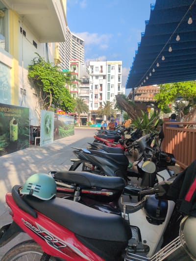 This photograph shows a street scene in South Quy Nhon Ward, Vietnam, with several parked motorbikes lining the sidewalk. The perspective is from ground level, looking down the street, and the image is rotated counter-clockwise. A blue awning hangs over a building on the left side of the frame, with lights strung underneath. Various promotional signs are displayed on the sidewalk next to the parked motorbikes. Buildings of varying sizes and architectural styles are visible in the background. Trees and foliage add touches of greenery throughout the scene. The foreground contains parked motorbikes, some with helmets resting on the seats. The presence of signage in Vietnamese script suggests the location is Vietnam.