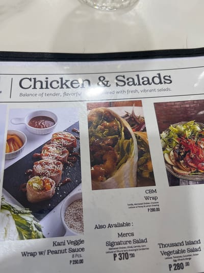 The image shows a menu featuring chicken and salad dishes. The menu items are displayed with photographs and descriptions. Featured items include "Kani Veggie Wrap w/ Peanut Sauce" priced at P250.00, "CBM Wrap" for P280.00, "Merce Signature Salad" at P370.00, and "Thousand Island Vegetable Salad" for P280.00. The menu is likely from a restaurant in Koronadal City, Philippines, as indicated by the Philippine Peso currency (P). The environment appears to be indoors, possibly a restaurant setting, with a white tablecloth and some natural light.