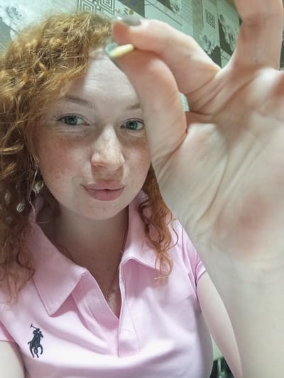 A young woman is depicted in a close-up, high-angle shot, likely lying down and looking upwards towards the camera. She has distinct red, curly hair, fair skin, and numerous freckles across her face. Her eyes are a light, striking green or blue-green, and she sports a gentle, relaxed smile.
She is wearing a light pink, collared shirt with the black Ralph Lauren polo logo (a polo player on a horse) embroidered on the left side of the chest. A thin, delicate metallic necklace is visible around her neck. Her right hand is extended prominently into the foreground, slightly out of focus, with fingers spread. Faint traces of light-colored nail polish (yellow and gray) can be seen on some of her fingernails.
The background on the right side of the frame shows a light-colored, patterned textile, possibly a bedspread or tablecloth, with a combination of geometric and floral designs. The lighting appears soft and even, typical of an indoor setting.
The woman's gaze is directed straight at the viewer, conveying a calm and friendly emotion. The only visible text is the Ralph Lauren logo on her shirt. The setting appears to be an interior space, without specific cues to indicate the time of day or weather beyond indoor conditions. The location context for this image is Yurgamysh, Russia.