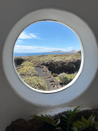 A bright coastal landscape is viewed through a circular, white-framed window. The perspective is from an interior space, with a section of a white, textured wall surrounding the opening. To the immediate right of the window, a green plant with several leaves grows, integrated into the interior wall or a nearby planter.

Beyond the circular frame, the scene reveals a clear, bright blue sky with delicate, wispy white clouds in the upper portion. Below the sky, a calm, deep blue body of water, likely the ocean, stretches horizontally towards the horizon. The middle and lower sections of the view are dominated by a rugged, dark, volcanic-looking coastline or cliff face. The terrain is characterized by dark, rocky textures and sparse, resilient green vegetation, appearing as low-lying shrubs or succulent-like plants clinging to the rocks. In the far distance, faint outlines of land formations, possibly mountains or hills, are visible above the sea.

The image suggests a sunny and clear day, likely midday or early afternoon, given the strong illumination. The volcanic rock and type of vegetation are strong indicators of a location like Lanzarote, aligning with the Haría, Spain context. No specific activity or event is taking place; it is a static scenic view. There is no visible text.