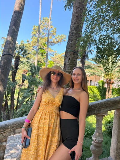 Two women stand close together outdoors. One woman, wearing a yellow patterned dress and a straw hat with sunglasses, holds a cell phone and wallet. The other woman wears a black strapless top and black shorts. They stand next to a stone railing. Palm trees and other greenery are visible in the background. Based on the architecture and landscape, the location appears to be in Bunyola, Spain.
