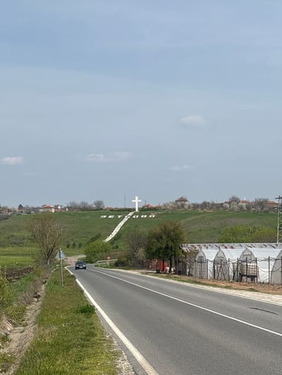 The image presents an outdoor scene in Zetiovo, Bulgaria, featuring a road that runs alongside a grassy field. In the field, a large white cross stands prominently. Adjacent to the road on the right, there are greenhouse-like structures. A car is positioned on the roadside. The sky is overcast.