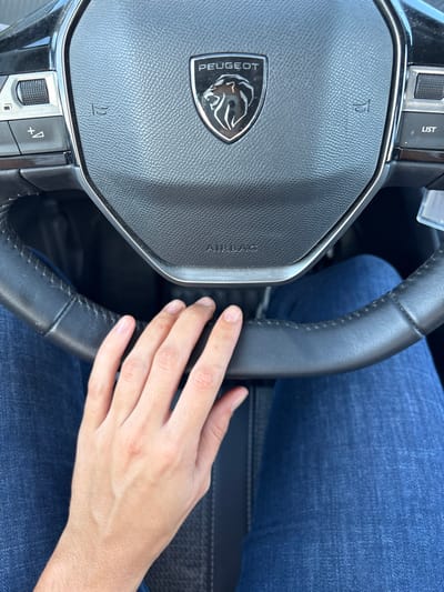 The image depicts the interior of a vehicle, focused on a steering wheel and the driver's lap. A black, leather-wrapped steering wheel occupies the central and upper portion of the frame. The steering wheel features the Peugeot manufacturer's emblem—a profiled lion's head with the word "PEUGEOT" above it—at its center. Below the emblem, the word "AIRBAG" is visible. Control buttons are present on both the left and right spokes of the steering wheel. A light-skinned human hand rests on the lower-left section of the steering wheel, with fingers curved around the rim. The individual's legs are visible below the steering wheel, clad in blue denim jeans. The lighting suggests an illuminated environment, possibly daylight. The specific location within the city of Villarrobledo, Spain, cannot be determined from the visual information provided in the image.