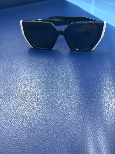 A pair of black-lensed sunglasses with thick white rims rests on a textured blue surface. The sunglasses are positioned on the left side of the frame, angled diagonally from the top left corner towards the bottom right. The blue surface appears to be leather or a similar material, characterized by a consistent, fine-grained texture. The lighting suggests it is daytime, with a bright light source casting subtle shadows and highlighting the texture of the blue surface. There are no people visible in the image. The scene is simple and focused on the object and its immediate surroundings. Some text is faintly visible on the inside of the sunglasses, appearing to be "PRADA". The context of Orsk, Russia, and any specific time of day or weather conditions are not directly inferable from the visual information in the image alone.