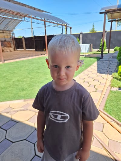 A young boy with blonde hair and a light-colored flower tucked behind his ear stands outside. He is wearing a dark gray t-shirt with a white oval logo and gray shorts. The boy is looking directly at the camera with a neutral expression. The background features a paved walkway, green grass, and a covered structure. The scene appears to be a residential outdoor area on a sunny day. No specific location in the city of Kara-Balta, Kyrgyzstan, can be identified from the image.