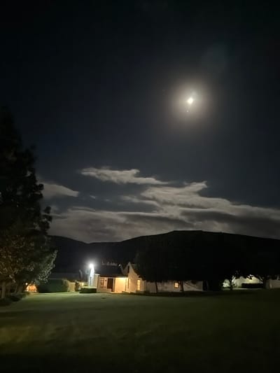 This vertical nighttime photograph captures a serene landscape illuminated by moonlight and artificial light sources. The upper portion of the image features a dark, expansive night sky. A brilliant, almost overexposed moon dominates the upper right quadrant, surrounded by a strong halo and subtle lens flare, suggesting its intensity. Near the moon, a distinct, smaller bright point is visible, possibly a star or planet. A prominent, elongated band of white clouds stretches horizontally across the mid-sky, intricately textured and brightly illuminated by the moon, creating a striking contrast against the dark sky.
The lower half of the frame presents a dark foreground with elements of a built environment and natural foliage. Dark silhouettes of trees and dense bushes are visible across the bottom and left edges. Further into the mid-ground on the left, several light-colored buildings, resembling houses, are partially visible, nestled among the trees. These structures feature lit windows and external lights, including a notably bright artificial light source near one of the buildings, casting light onto its immediate surroundings. The ground in the foreground is dark, likely a grassy area.
No people are visible in the image, nor is there any discernible activity beyond the static scene. The time of day is clearly night, with fair weather allowing for clear visibility of the moon and stars, alongside distinct cloud formations. The artificial lights provide a sense of human presence in an otherwise natural and tranquil setting. There is no visible text. The image provides a peaceful nocturnal view within a residential or semi-rural area in the Overstrand Local Municipality, South Africa.