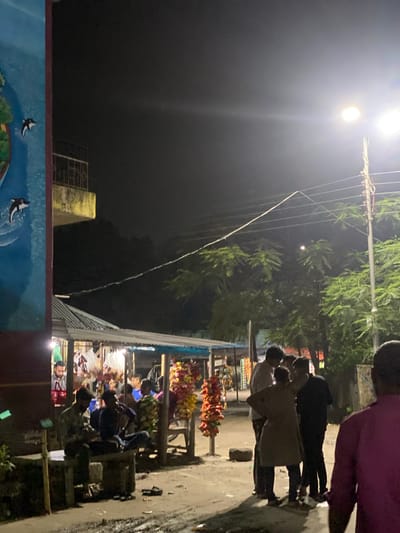 The image depicts an outdoor scene at night in Tarabo, Bangladesh. A street market is illuminated by a bright overhead light. Several people are visible, some standing and browsing at stalls displaying various items, including what appears to be snack foods. Tables are set up with goods displayed on them. The ground is wet, possibly from a recent rain. Overhead power lines stretch across the scene. A painted mural with aquatic imagery is visible in the foreground.