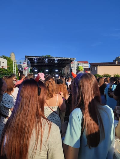 The image captures a crowd of people gathered in front of a stage, likely at an outdoor event or concert. The sky is a clear, bright blue. In the foreground, the backs of several individuals are visible, their hair ranging in color from light brown to red. Some are wearing accessories, including devil horns. Behind them, a stage structure with lighting is set up, and a large bubble containing a Hello Kitty balloon is suspended to the left of the stage. Trees and buildings are visible in the background, suggesting an urban or semi-urban setting. The overall scene suggests a lively, festive atmosphere. This image is located in the city of Korosten, Ukraine.