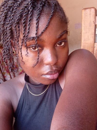 This image is a close-up portrait of a young adult female, taken in an indoor setting in the city of Ejule, Nigeria.

The subject has dark skin and dark, twisted braids, with several strands hanging across her forehead. Her dark eyes are accentuated with eyeliner, and she maintains a direct gaze towards the camera. Her lips are full with a light pinkish hue. She wears a black halter-neck top and is adorned with a thin gold-colored chain necklace. A small, gold-colored earring is visible on her left earlobe. Her right shoulder and upper arm are partially visible in the lower right foreground.

The background consists of two distinct areas. To the left, a patterned fabric, possibly a curtain or textile, with red and white floral or abstract designs is partially visible. To the right, a plain, light brown wall is present, behind a vertical wooden structure, possibly a door frame or furniture element. The scene is illuminated evenly, suggesting natural light.