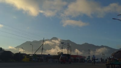 A bustling street scene unfolds beneath a hazy sky with scattered clouds. A mountain range shrouded in mist forms a dramatic backdrop. Utility poles and wires crisscross the foreground. A red three-wheeled vehicle moves along the road, alongside a cyclist. People gather near structures lining the street. The overall atmosphere suggests a vibrant, active community nestled in a scenic location.