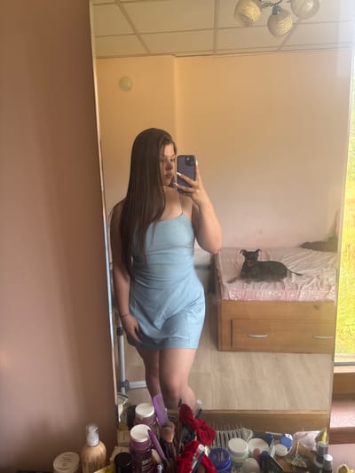 The image is a selfie taken in a room, reflected in a mirror. The primary subject is a young woman with long, dark hair, wearing a sparkly blue dress. She is holding a smartphone in her left hand and is posing for the camera. In the reflection, a small black dog is visible lying on a bed in the background. The room appears to be a bedroom or dressing area, with furniture and a collection of makeup and beauty products visible on shelves to the right of the woman. The lighting suggests it could be daytime, possibly indoors with natural light coming from a window. There are no specific indicators of the weather or time of day beyond that. The overall mood seems casual and self-aware, typical of a selfie. No visible text is legible in the image. The location context provided is Prodanovtsi, Bulgaria.