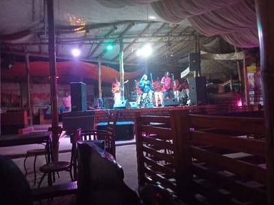 The image shows a live music performance at a venue in Bungoma, Kenya, located at latitude 0.593 and longitude 34.559. Five individuals are on stage, with one woman singing into a microphone and four men playing musical instruments.  The stage is illuminated by spotlights, and a large speaker is positioned on each side of the stage. The venue is a partially enclosed structure with a thatched roof and wooden walls. The space is sparsely furnished, with a few tables and chairs in the foreground. The image was taken from a seated perspective in the audience.