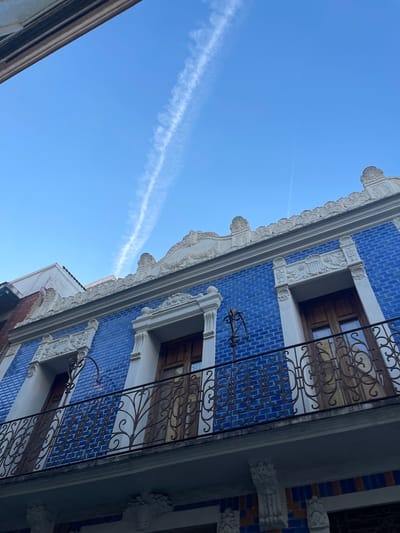 The image shows a low-angle view of a building facade in Paiporta, Spain. The facade is adorned with blue tiles, white architectural trim, and wrought iron balconies. The sky is clear blue with a visible contrail. The building has wooden-framed windows, and ornate decorative elements surround them. The image is taken from the street, looking upward at an angle.