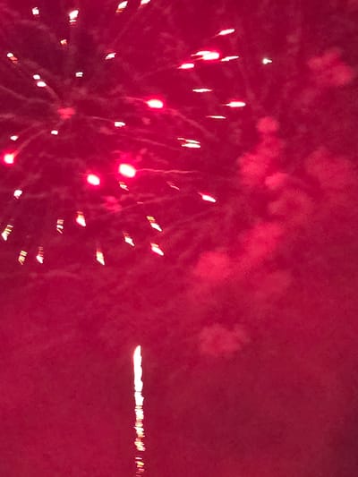 This is a photograph of a fireworks display against a dark, reddish-pink sky. The fireworks are predominantly red and white, with some appearing as bursts of light and others as streaks or trails. There are numerous small, bright points of light scattered across the upper portion of the image, suggesting multiple explosions or sparks. In the lower center of the image, a single, long, vertical streak of white light with red at its base suggests a firework that has just been launched or is in the process of descending. The overall impression is one of a nighttime celebration. The image does not provide any specific landmarks or details that would allow for precise localization to Johns Creek, United States of America.