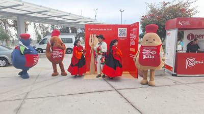 In Tacna, Peru, an outdoor event is taking place. Two mascots, one resembling a blueberry and the other a potato, stand in the foreground. The blueberry mascot wears a red apron and holds a white spoon, while the potato mascot also wears a red apron with the text "Feria Gastronómica Perú mucho gusto" and carries a white spoon. Behind them, three individuals are posing in front of a large red backdrop displaying "25 al 28 de julio de 2026" and a QR code. The backdrop also features graphics of a hand and Peruvian cultural motifs. To the right, a red kiosk with the "Prom Perú" logo and more "Perú mucho gusto" branding is visible. A person is working at a desk inside the kiosk. A white van and a car are parked in the background. The scene is set on a paved area with trees and a cloudy sky overhead.