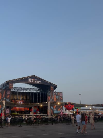 A wide-angle, eye-level shot captures an outdoor music festival stage set up in a parking lot at dusk in the Polígon Industrial De Son Castello, Spain. The stage, constructed with metal trussing and covered with a black canopy, displays the logo "SONO FUSTERET" in white lettering. A large screen behind the stage shows a Hyundai logo above "Proa Automocion" text, with a green car and a large, inflatable red cartoon character with sunglasses above it. The scene is decorated with colorful banners featuring geometric patterns and palm trees. In the foreground, a few individuals are walking and standing near scattered tables and chairs, set up on the asphalt. The sky is a clear, pale blue, transitioning to a lighter hue near the horizon.