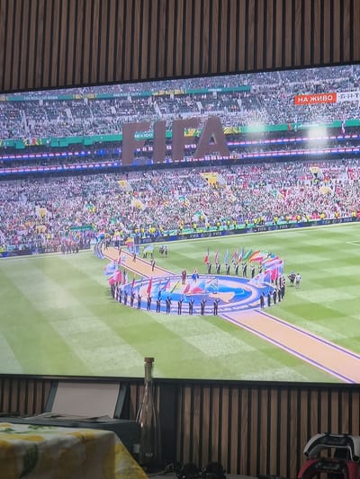 The image shows a large screen displaying a FIFA event, likely a stadium with a crowd and a ceremonial parade on the field. Large letters spelling "FIFA" are visible on the screen, and the text "НА ЖИВО" (meaning "LIVE" in Russian) is displayed in an orange box. The event appears to be a soccer match opening ceremony, with people in uniform holding flags of various countries. The setting appears to be indoors, with a wooden paneled wall in the background and a table with a bottle and what looks like a printer in the foreground. The image is taken from a viewing angle, suggesting the screen is part of a television or large display. The city of Xàtiva, Spain is not depicted in the image; the visual content solely pertains to a FIFA event shown on a screen.