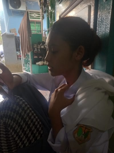 A young individual, shown in side profile facing left with eyes downcast or closed, is seated in what appears to be a semi-outdoor residential setting within the city of Bojongherang, Indonesia. The individual wears a white long-sleeved shirt, consistent with a school uniform, featuring a distinct emblem on the right sleeve with green and brown elements and the partially legible text "CIANJUR." Dark-colored lower garments are also visible. The individual's right hand is positioned near the collar of the white shirt. A black and white houndstooth-patterned bag rests in the lower-left foreground. Behind the subject, a dark green door frame is visible. The background to the left reveals a light blue exterior building wall with an outdoor air conditioning unit mounted high, below which a light brown piece of fabric hangs. A white decorative metal gate and a green-framed window are also present. A collection of footwear, including various sneakers and sandals, is arranged on a shoe rack on a light-colored ground surface. The scene is illuminated by natural daylight.