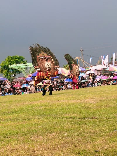 Two people are performing a traditional dance in the city of Karangmalang, Indonesia. They are wearing large, colorful costumes with large, exaggerated animal heads, possibly representing lions. The dancers are standing on a grassy field with a large crowd of people watching them. There are many umbrellas and other colorful items in the background. The sky is grey and overcast. The scene is likely a cultural celebration or a festival. The performance is taking place on a field surrounded by a dense crowd of people, all looking at the dancers. The performers are walking toward the crowd, with their costumes and headpieces being very elaborate and detailed. There are several vendor stalls and stalls set up in the background. The scene is lively, vibrant and celebratory. The location is likely a field or open area in a public space in the city of Karangmalang, Indonesia. The coordinates -7.47, 111.064 pinpoint the location of the image as being in Karangmalang, Indonesia.