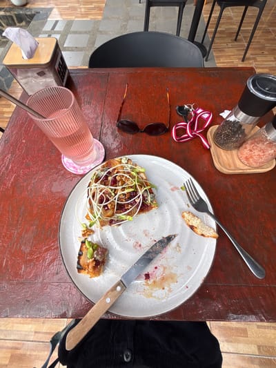 An overhead view depicts a reddish-brown wooden table in an outdoor or semi-outdoor setting in Koh Tao, Thailand; the specific establishment is not discernible from the image. A partially consumed meal occupies a light-colored ceramic plate with a darker rim. The central food item is a slice of toast topped with a dark red spread, blue cheese crumbles, walnuts, and green sprouts. A smaller, severed portion of this dish is positioned on the lower left of the plate, adorned with green pistachios. A small, plain piece of toasted bread lies on the upper right. A metal fork is on the right side of the plate, and a serrated knife with a light wooden handle rests on the lower center, amidst scattered food remnants and a red liquid residue. To the upper left, a wooden-topped tissue box containing white tissues is present. Adjacent to the plate on the left is a tall, ribbed, pink-tinted glass containing a light pink liquid and a metal straw, situated on a round, pink marble-patterned coaster. To the right of the glass, folded tortoiseshell sunglasses and a set of keys on a red and white striped lanyard are placed on the table. On the far right, a small wooden tray holds two clear cylindrical grinders, one filled with black peppercorns and the other with pink Himalayan salt. The background features a tiled floor with alternating light grey and darker grey square tiles, along with portions of dark, minimalist chairs. The lap of an individual, dressed in dark clothing, is visible at the bottom of the frame, indicating a seated person.