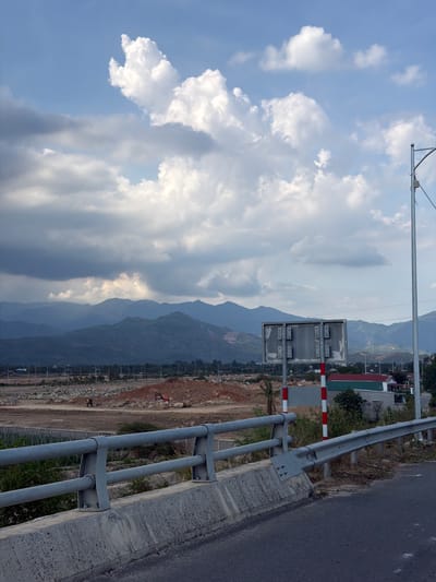 A roadside scene in an unidentified location in Vietnam. A gray, metal guardrail runs along the right side of the frame, adjacent to a gray asphalt road. Beyond the guardrail, the terrain is largely barren, with patches of brown dirt and scattered vegetation. In the distance, rolling mountains fade into a hazy blue. A large bank of white clouds fills the left side of the frame, contrasting with patches of blue sky. Above the guardrail, a utility pole extends horizontally across the frame. A sign with red and white stripes is visible along the roadside.