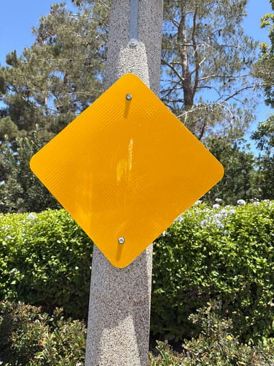 A bright yellow, diamond-shaped, reflective sign is mounted on a rough, speckled, grey concrete pole. The sign is tilted at an angle and has two silver bolts securing it to the pole. A faint, vertical white streak is visible on the sign's surface. Behind the pole and sign, a dense green hedge with small white flowers is prominent. Beyond the hedge, a tall pine tree with brown bark and green needles is visible against a clear blue sky. The overall scene suggests an outdoor environment, possibly a park or roadside in Aliso Viejo, California, United States of America.