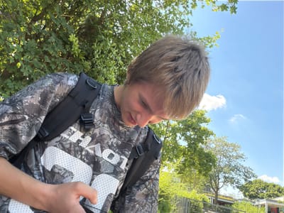 The image shows a person with blonde hair wearing a camouflage-patterned shirt and a black backpack. The person is looking upwards, and their face is partially visible. The background consists of green trees and a bright blue sky with a few white clouds, suggesting it is daytime and the weather is fair. The perspective of the photo is from below, looking up at the person and the sky. There is no discernible activity taking place beyond the person being present in the scene. The image appears to be taken outdoors in a natural environment with trees. The text visible on the person's shirt is white and appears to be letters, possibly part of a brand name or logo, but it is not entirely legible due to the camouflage pattern and the angle. No specific location cues beyond the presence of trees and sky are visible, although the prompt provides the location context of Dorsten, Germany. The lighting suggests it is likely daytime, possibly midday given the brightness of the sky.