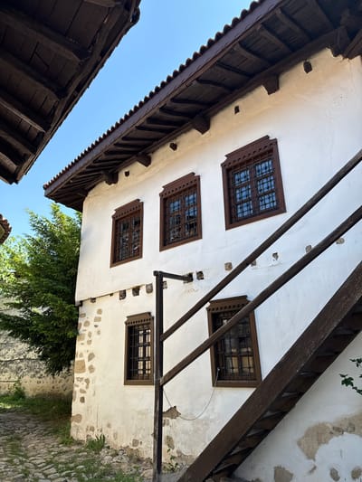 The image displays a traditional two-story building in Sandanski, Bulgaria, characterized by white plastered walls and dark wooden architectural elements under a bright blue sky. The upper floor features three prominent rectangular windows with ornate dark brown wooden frames and intricate black metal grilles. Beneath the roof, dark exposed wooden beams and rafters are visible, supporting terracotta roof tiles. The lower section of the building's facade shows patches of rough-hewn stone masonry interspersed with the white plaster. Two additional windows with similar dark wooden frames and grilles are visible on the lower floor.
An exterior, dark brown wooden staircase ascends diagonally from the lower right to the mid-right, leaning against the white wall. To the left of the building, a dense green tree or bush provides a natural backdrop. The ground in the lower left consists of a cobblestone path with sparse grass and small plants growing between the stones and at the base of the wall. In the upper left quadrant, another dark wooden roof structure is partially visible, framing a section of the clear blue sky. No individuals or animals are present in the scene. The lighting suggests a sunny day, with clear shadows.