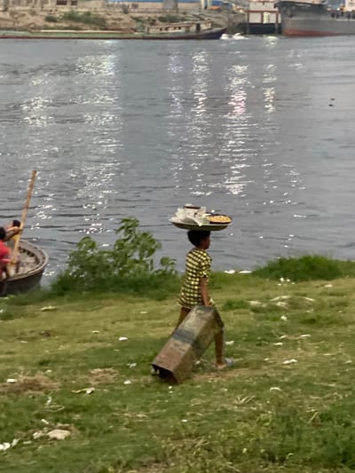 This image captures a tranquil riverside scene in Tarabo, Bangladesh, likely during the daytime. A wide body of water, possibly a river or canal, fills the left side of the frame, its surface shimmering with reflections of light, suggesting either sunlight or artificial light sources reflecting off the ripples.
On the right, a lush green grassy bank runs alongside the water. A person, dressed in a light-colored, possibly checkered, shirt, is depicted walking along this bank. They demonstrate traditional carrying methods, balancing a round, flat woven basket or tray on their head. Simultaneously, they hold a large, rectangular, and somewhat worn container, possibly made of metal or wood, which they appear to be either carrying or dragging along the ground. The individual's features are not distinct enough to discern specific emotions.
Further into the water on the right, a small, dark-colored boat is partially visible. At least one person, and possibly two, are seated or standing within the boat, with one person holding a long pole or oar, suggesting navigation or propulsion.
In the distant background, on the far side of the water, faint outlines of buildings and structures can be seen, hinting at a settlement or urban area across the river. The overall scene portrays a slice of everyday life and activity along a waterway. There is no visible text in the image.