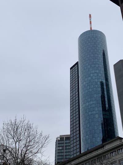 The image shows a view of the cityscape of Frankfurt am Main, Germany, specifically the skyscraper complex known as the "Twin Towers" located at coordinates 50.11° N, 8.669° E. The view is taken from the ground level, looking upward at the two main towers, with a slightly overcast sky above. The dominant tower is the "Tower 185", with a red and white antenna protruding from the top. The taller of the two, it has a cylindrical shape and is fully clad in glass. The reflection of a nearby building is visible in the glass facade. To the left, partially obscured by the "Tower 185", is the "Tower 120". Both buildings have a dark-toned window frame and are constructed from glass and metal. In the foreground, a branch of a bare-branched tree partially obstructs the view of the buildings. At the base of the "Tower 185", there is a building with a flat roof and visible balconies, potentially residential or commercial. The image captures the city's modern architectural landscape and showcases the prominent role of skyscrapers in Frankfurt's skyline.