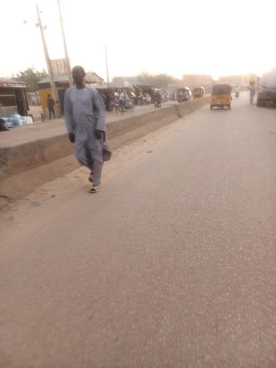 The image captures a bustling street scene in what appears to be a semi-urban or urban area, identified by the context as Tudun Murtala, Nigeria. In the foreground, a man in a light gray traditional garment, possibly a kaftan or djellaba, walks along the edge of a paved road towards the left. He is wearing sandals.
The road occupies the center and right side of the frame, showing light wear and a slightly dusty appearance. On the left side of the road, a concrete barrier separates the main thoroughfare from a lower section where shops or stalls are set up. Numerous people are visible in the mid-ground on this left side, some gathered around the stalls, others appearing to be walking or riding motorcycles. Several motorcycles are parked or moving slowly. Further down the road on the right, multiple yellow auto-rickshaws, commonly known as Keke Napep in Nigeria, are visible, suggesting active traffic. A larger vehicle, possibly a truck, is partially seen on the far right. Utility poles line the left side of the road, indicating infrastructure.
The lighting suggests late afternoon or early morning, with a soft, warm, and somewhat hazy glow across the sky and scene. The atmosphere appears calm but active, depicting everyday life in the area. There is no visible text in the image.