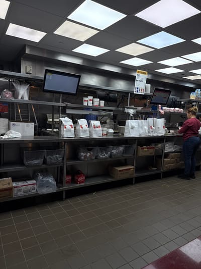 A restaurant setting shows a worker standing behind a long stainless steel counter filled with packaged food and condiments. Two monitors are mounted on the counter, one displaying menu items. Above, a diamond-patterned ceiling provides bright lighting. The floor is tiled. The scene suggests a fast-food or casual dining environment.