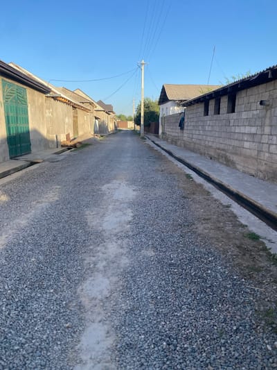 The image shows a gravel road in a residential area, likely in the city of Боғистон, Tajikistan. The road is lined with buildings on both sides. On the left, there are several single-story structures with plastered walls and pitched roofs. One building features a large, ornate green metal gate. On the right, there is a longer, one-story building constructed of grey concrete blocks with an open, roofed section. A dark, narrow drainage ditch runs along the edge of the road in front of this building. Overhead power lines stretch across the clear blue sky. The overall impression is of a somewhat unpaved, rural or semi-urban street.
