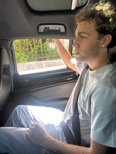 The image, rotated 90 degrees clockwise from a standard upright view, captures a man leaning into the interior of a car during what appears to be a bright, sunny day in Torres Vedras, Portugal.
The man, with light skin, curly brown hair, and a short beard, is the primary subject. He is wearing a light blue polo shirt and light blue trousers. His head is tilted downwards and to the right within the frame, and his gaze is directed intently at something out of view below him, suggesting concentration or observation. A black seatbelt is visible across his chest and lap. His right hand is clenched and resting near his leg.
The setting is inside a car, with elements of the dark gray/black interior visible, including the headliner, an A-pillar with small control buttons (possibly for garage door openers or interior lights), and part of the dashboard with an air vent. Through the car window, an outdoor scene is visible, featuring lush green foliage (likely trees or bushes) and a light-colored fence or lattice structure, indicative of a garden or residential area.
Bright natural sunlight streams into the car, creating strong highlights on the man's hair and clothing, and producing some glare within the vehicle. There is no visible text in the image.
