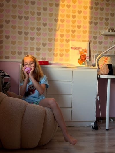 The image shows a young girl sitting on a plush, cream-colored armchair in a room with light brown wooden flooring. She is wearing a light blue t-shirt with a cat print and has blonde hair and glasses. She is holding a round, pink object up to her face, appearing to interact with it. Her left leg is extended, and her right leg is bent.
The background features a wall with a heart-patterned wallpaper in shades of pink, brown, and cream. To the left of the armchair, a white unit is visible with a book featuring an illustration of a pumpkin and flowers. Above this unit, a lamp and a pink salt lamp are present. The scene appears to be indoors, likely a bedroom, and the lighting suggests it could be daytime, possibly late afternoon due to the warm glow. There is visible text on the book cover, which reads "Fruits And Flowers". The overall mood is casual and playful.