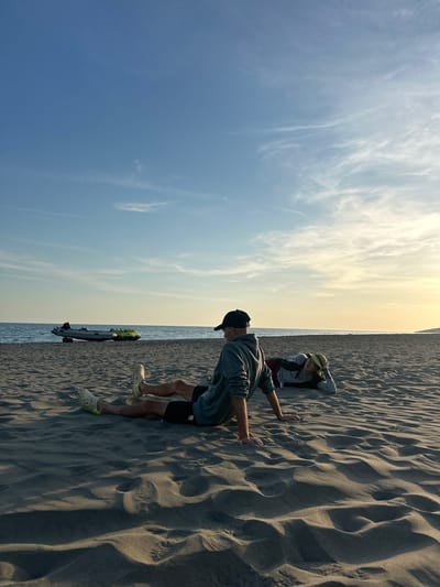 The image captures a scene on Velika Plaža (Long Beach) in Donji Štoj, Montenegro. The sky transitions from a pale blue to a faint yellow near the horizon, marked by wispy clouds. Below the sky, a strip of water is visible, indicating the Adriatic Sea.
On the sandy beach, two people are present. A man wearing a dark baseball cap, a gray sweatshirt, black shorts, and light-colored Crocs with yellow accents stands with his hands on the sand, leaning forward. Behind him, a person wearing a hat and a long-sleeved shirt is partially obscured in the sand. A sandboard with visible yellow and gray elements sits near the edge of the water. The sand itself is heavily textured, with prominent wave-like formations and visible footprints.