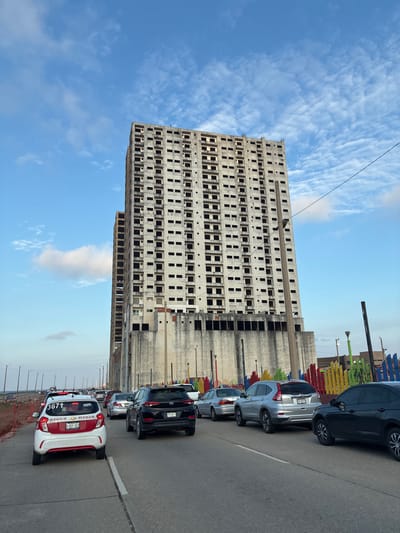 A vertical, wide shot showcases an unfinished concrete building against a blue, partially cloudy sky. The building features multiple rows of empty window spaces, a stark contrast to its gray, unadorned facade. To the right, cars are parked along a gray asphalt road. A red-and-white taxi with the license plate "A-431-XEY", bearing the number "3671" and the text "LOC DE COATZA VER" is in the foreground. The license plate "XZW-261-C" is visible on the black car behind. The location is Coatzacoalcos, Mexico.