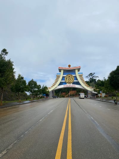 A forward-facing, eye-level, medium shot captures the Bo Y International Border Gate in Bo Y, Vietnam. The gate, an imposing structure with curved, tiered architecture and a central circular emblem featuring Vietnamese script and symbols, is the focal point of the image. The roadway leading to it is wet, reflecting the overcast sky, and marked with double yellow lines. On the left side of the road, a gently sloping hill is covered in greenery and trees. On the right, a line of smaller trees separates the road from the border control area, where a white bus and several individuals are visible. The sky above is a muted, cloudy grey.