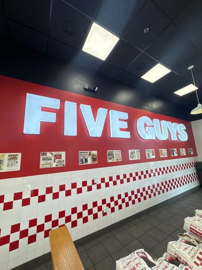 This image shows the interior of a Five Guys restaurant. The restaurant's logo, "FIVE GUYS," is prominently displayed in large, illuminated white letters on a red wall. Newspaper clippings and magazine articles are pinned to the wall beside the logo. The floor has a red and white checkered pattern. To the right of the floor is a gray tiled wall with brown bags hanging on it. The black ceiling above has square, recessed lights. There is no indication of its location in Bloomingdale, United States of America.