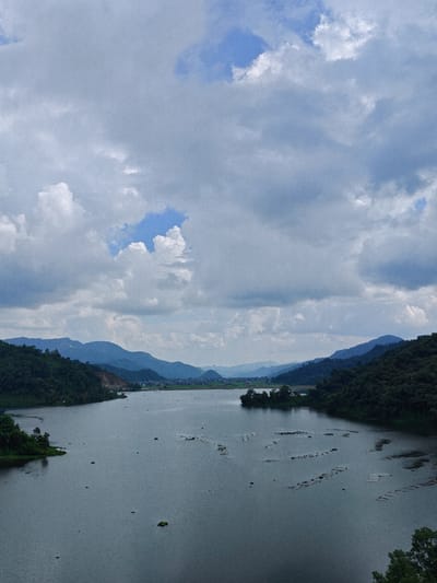 The image shows a wide, calm lake with gentle ripples. Dense green trees and rolling hills frame the left and right sides of the lake, with a series of distant, muted blue mountains forming the horizon. The sky is overcast with large, fluffy white clouds interspersed with patches of blue. Scattered across the surface of the lake are clusters of what appear to be floating vegetation or small islands. In the far distance, a small settlement with buildings can be seen nestled between the hills. The overall impression is one of a serene, natural landscape under a cloudy sky. This scene is characteristic of the Begnas Lake area in Lekhnath, Nepal, known for its natural beauty and mountainous surroundings.