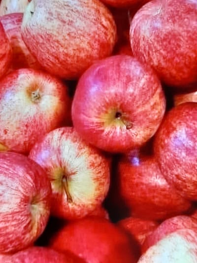 The image displays a close-up, overhead view of a pile of ripe apples. The apples are predominantly red with some yellow and white marbling. Their surfaces appear slightly textured, with some showing subtle imperfections. The lighting is warm, highlighting the vibrant colors of the fruit. The arrangement suggests they are being displayed or stored together. There is no indication of the city of Voluntari, Romania, in the image.