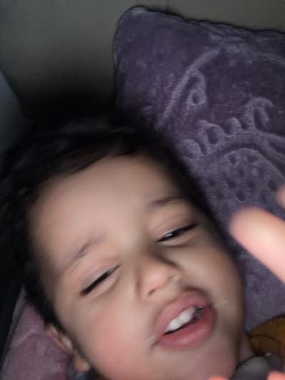 This image depicts a young child, possibly a toddler or preschooler, captured in an indoor setting in Dar Bouazza, Morocco. The child, with dark hair, light-to-medium skin, and an unclear gender, is positioned centrally in the frame. Their eyes are partially closed or squinting, and their mouth is slightly open, revealing several upper and lower teeth. A whitish substance is visible around the lips, suggesting food or drink residue. A blurred finger, likely belonging to the child or another individual, is present in the foreground on the right, partially obscuring the child's face. The child appears to be lying down on a soft surface, possibly a bed or sofa. The background consists of a dark purple, textured blanket or pillow with an indistinct pattern, visible to the upper right and behind the child's head. A darker, unidentifiable area is visible to the upper left. The lighting is dim, and the overall image exhibits slight motion blur, particularly in the background and foreground finger.