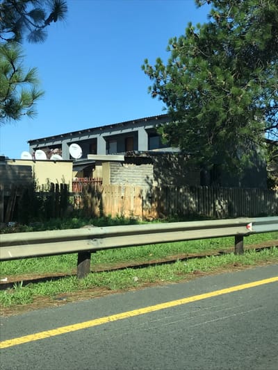 A two-story house stands on the roadside under a clear blue sky. The house's architecture includes brick and corrugated iron elements. Satellite dishes are mounted on the building's facade. A lush green tree partially obscures the view of the house. A grey guardrail divides the house from a paved road with a yellow line marking. The road is next to the shoulder with grass.