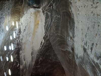 The image captures an interior view of the Turda Salt Mine in Turda, Romania, showcasing its unique geological formations and artificial lighting. The scene is dominated by the rough, textured walls of the salt mine, which exhibit layered striations of varying colors, including dark browns, grays, and whites, suggesting compressed salt deposits. Areas of white, crystalline salt formations are prominently visible, some appearing powdery and others more consolidated.
Suspended by thin wires, a series of modern, cylindrical fluorescent lights illuminate the cavernous space. These lights are arranged in vertical lines and emit a bright, white glow, creating distinct reflections on the salt surfaces. The lighting casts shadows that accentuate the uneven topography of the walls and the texture of the salt.
A dark, vertical cable or pipe runs down the center of the frame, further breaking up the natural contours of the salt walls. Fine netting or mesh is visible in some sections, likely installed for structural support or safety, adding another layer of texture to the environment. The overall impression is one of an otherworldly, subterranean landscape, artificially enhanced by man-made illumination.