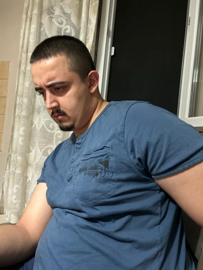 A close-up, vertically oriented photo shows a young man in a blue t-shirt. He has short dark hair, a neatly trimmed beard, and is looking down and to his left. His right arm is bent and resting against his side, while his left arm is out of frame. The background consists of a window with a black screen or blind and sheer white curtains. There is some text visible on the t-shirt, though it is partially obscured and difficult to read clearly. The overall impression is of a casual indoor setting, possibly at dusk or night due to the dark window. No specific activity or event is clearly depicted, and the man's expression is neutral or slightly pensive. The location context of Byala Slatina, Bulgaria is not visually evident in the image itself.