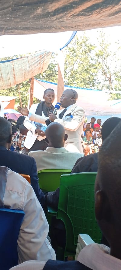 Two men stand before a seated audience under a partially covered structure in Kalemie, Democratic Republic of the Congo. The man on the right holds a blue microphone and speaks, wearing a light-colored suit. The man on the left holds a water bottle and wears a white jacket over a black shirt. The audience consists of men, women, and children seated in plastic chairs of varying colors. Trees and the sky are visible in the background.